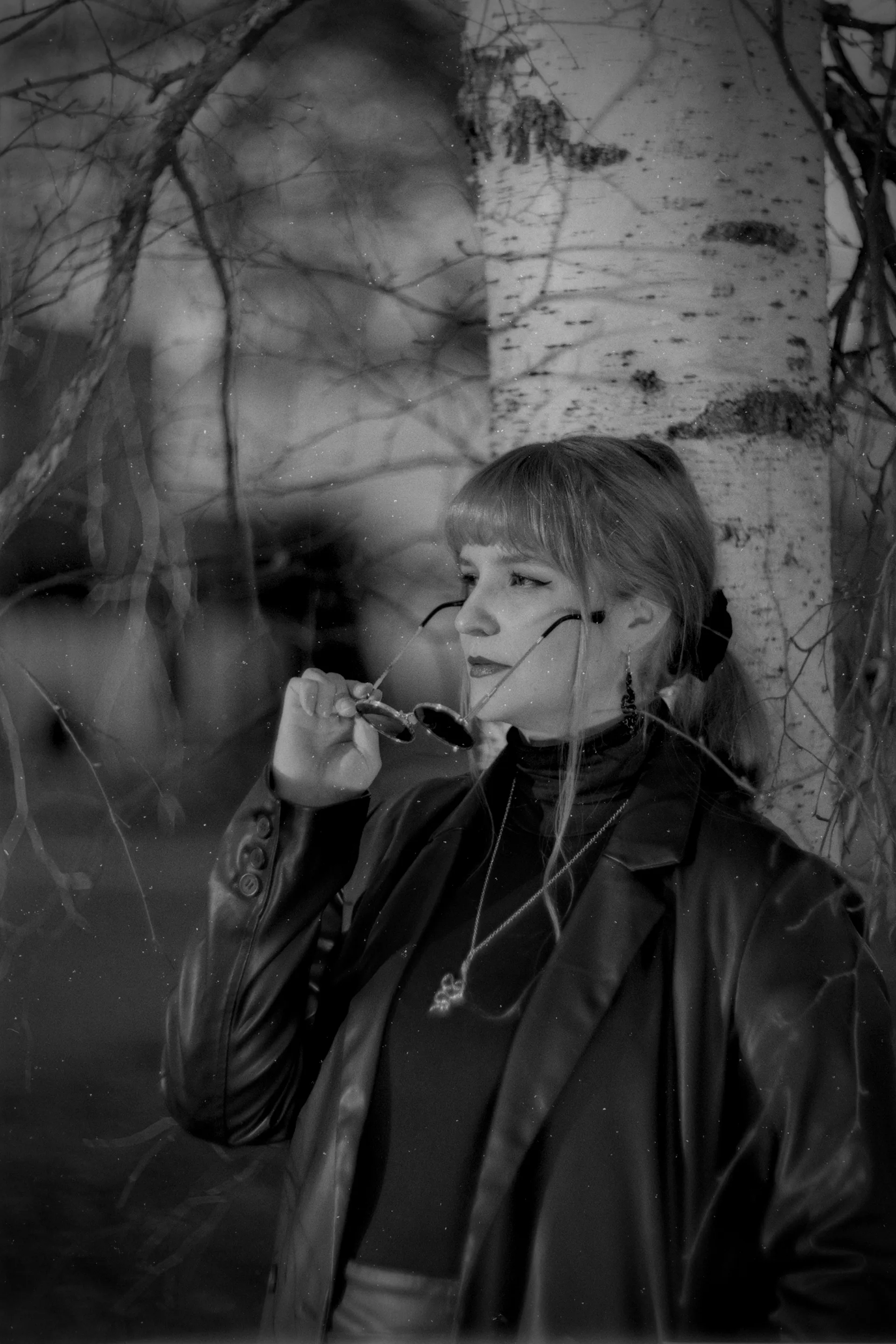 Mustavalkoinen filmikuva naisesta koivun vieressä aurinkolasit kädessään.